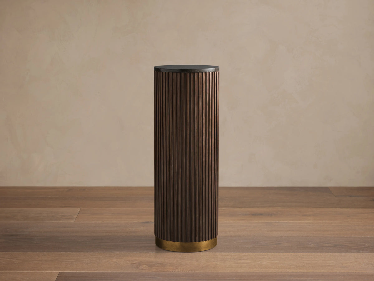 Finnley Pedestal table with dark ribbed wood finish, cylindrical shape, and brass base, set on a wooden floor against a beige wall.