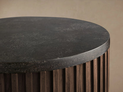 Finnley Pedestal table with dark ribbed wood finish, cylindrical shape, and brass base, set on a wooden floor against a beige wall.