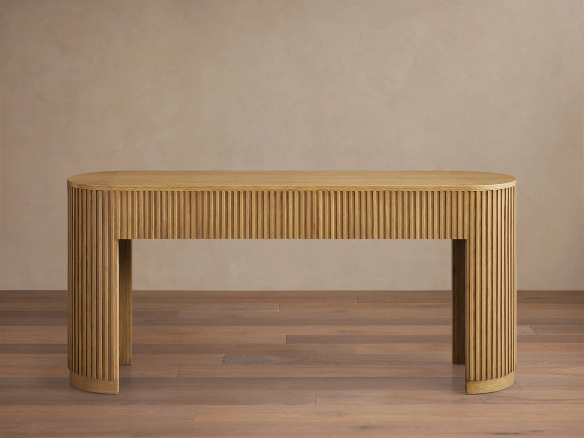 Finnley Wood Console Table with a Honey finish, features rounded edges and vertical grooves, set on a wooden floor against a beige wall.