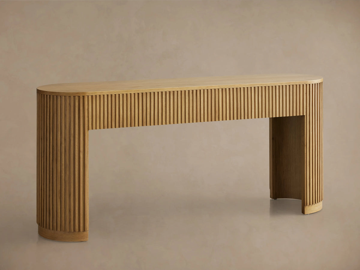 Finnley Wood Console Table with a Honey finish, features rounded edges and vertical grooves, set on a wooden floor against a beige wall.
