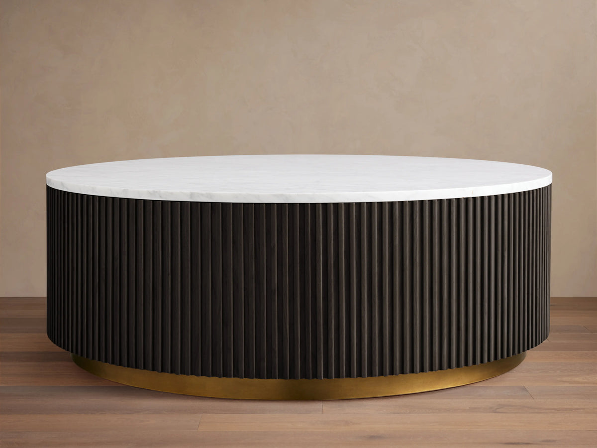Finnley Round Coffee Table with a white marble top, dark ribbed cylindrical base, and brass-finished plinth on wooden floor.