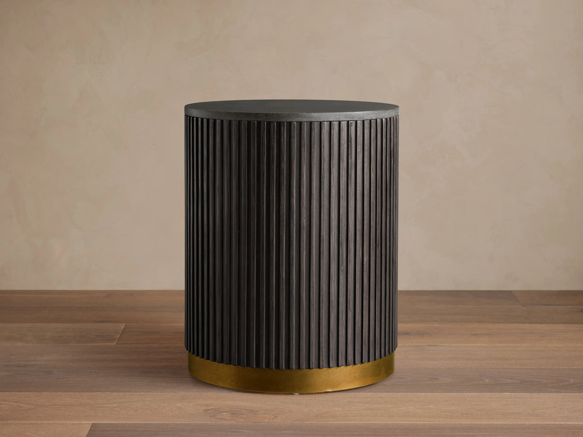 Finnley End Table with vertically ribbed dark wood surface, brass-finished base, standing on a wooden floor against a beige wall.