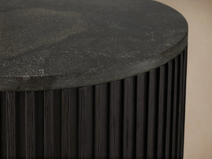 Finnley End Table with vertically ribbed dark wood surface, brass-finished base, standing on a wooden floor against a beige wall.