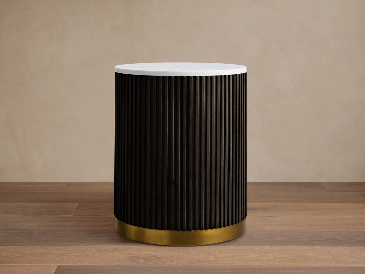 Finnley End Table with vertical dark ribbed sides, white round top, and brass-finished base on a wooden floor.