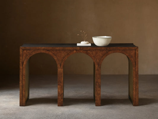 Hattie Burl Console Table: brown burl wood with three arches, dark top, neutral bowl & small decor items on top, against a beige wall.