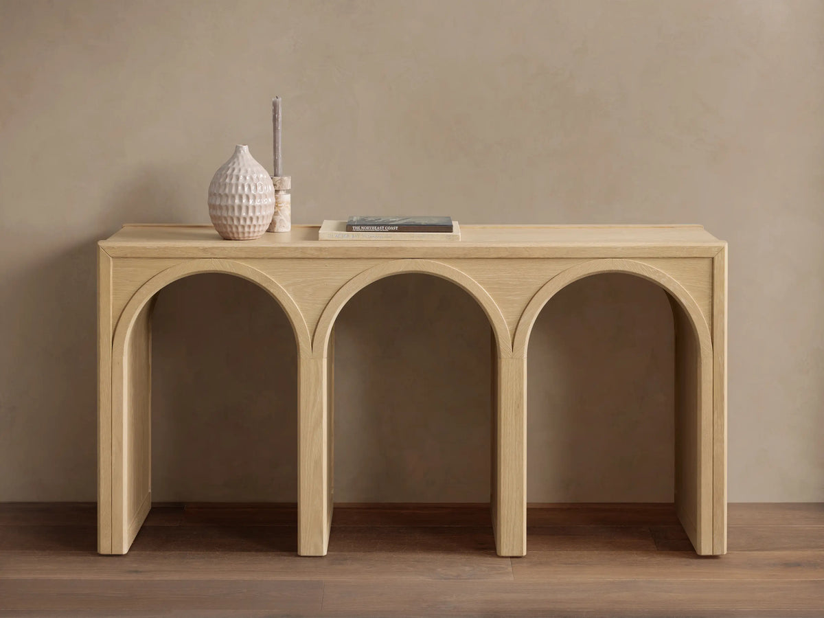 Oak Hattie Console Table with three arch-shaped supports, light wood finish, vase, book, and candle displayed on top.