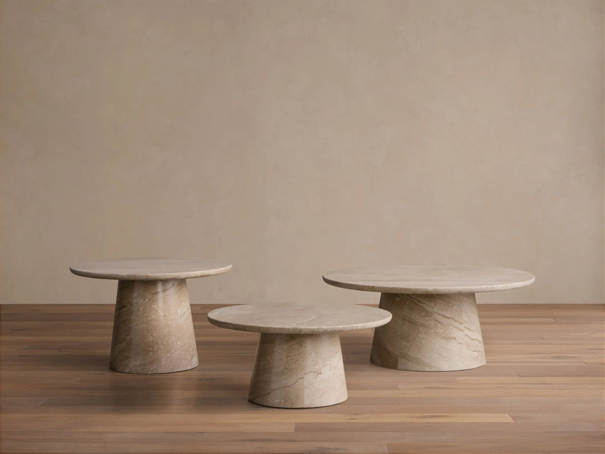 Three round beige marble Hemmings Nesting Coffee Tables of varying heights on a wooden floor against a beige wall.