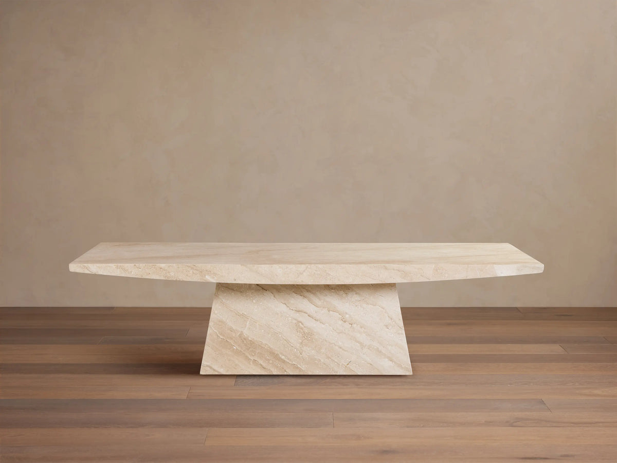 A beige marble table with a rectangular top and an angled, trapezoidal base. Positioned on a wooden floor against a plain wall.