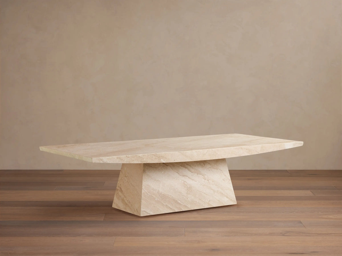 A beige marble table with a rectangular top and an angled, trapezoidal base. Positioned on a wooden floor against a plain wall.