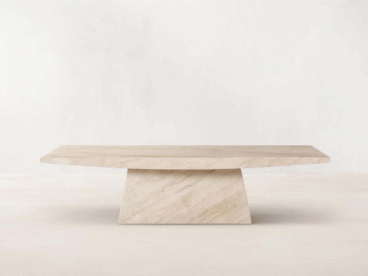 A beige marble table with a rectangular top and an angled, trapezoidal base. Positioned on a wooden floor against a plain wall.