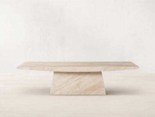 A beige marble table with a rectangular top and an angled, trapezoidal base. Positioned on a wooden floor against a plain wall.