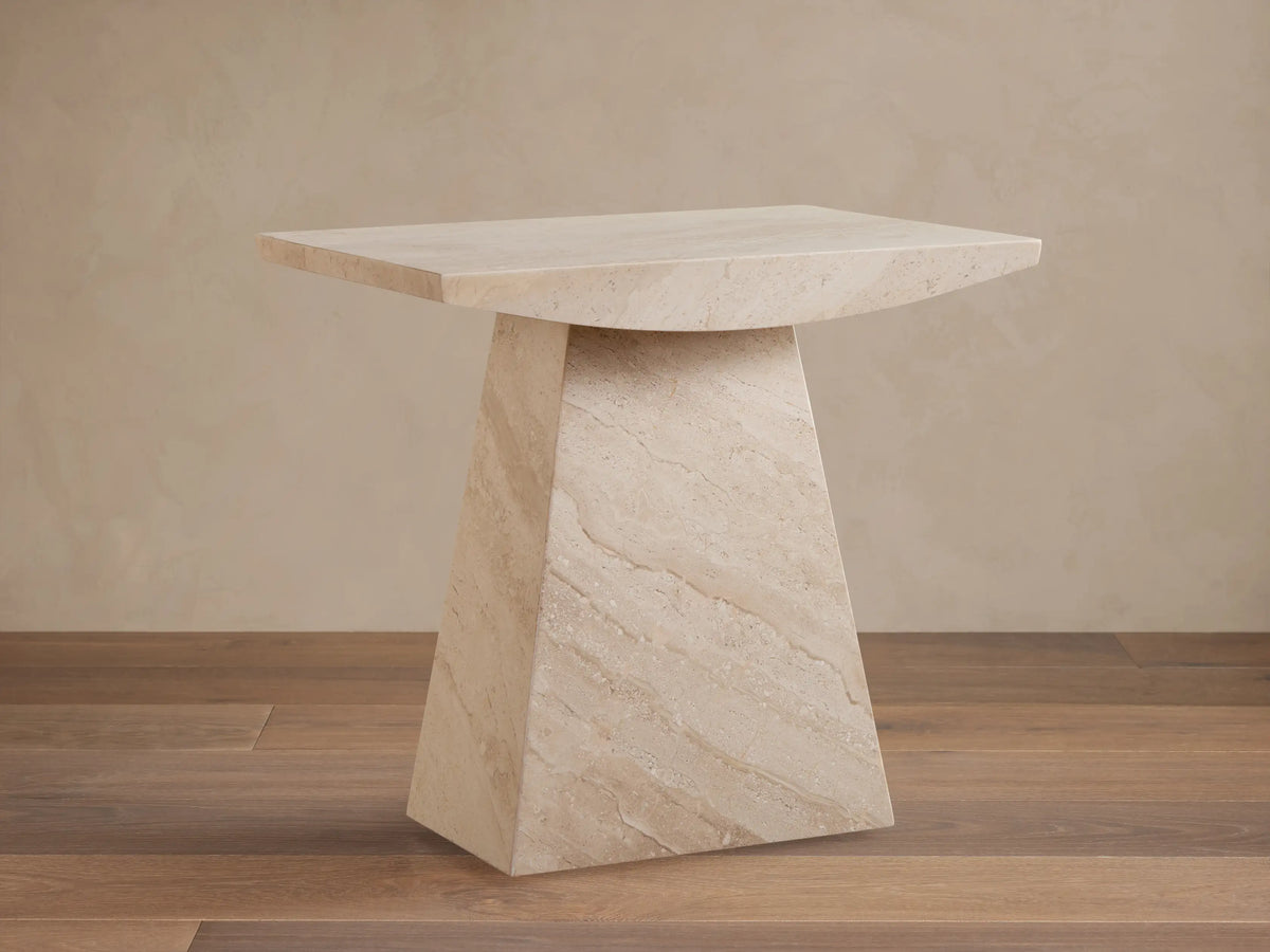 A beige marble side table with a trapezoidal base and a rectangular top, set on a wooden floor against a light wall.