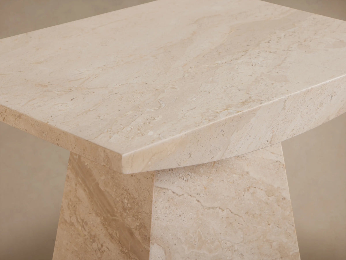 A beige, rectangular stone table with a narrow base and wider top, set on a wooden floor against a neutral wall.