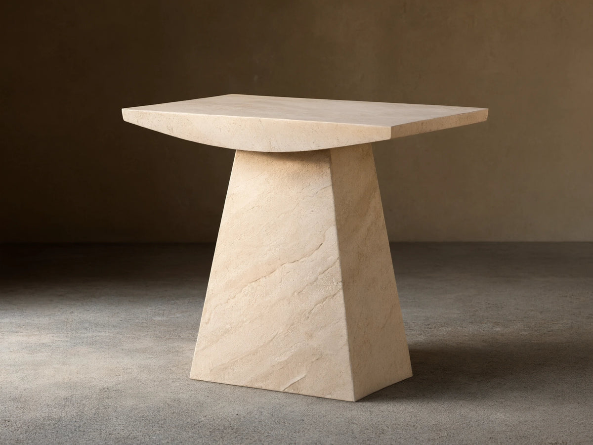 A beige marble side table with a trapezoidal base and a rectangular top, set on a wooden floor against a light wall.