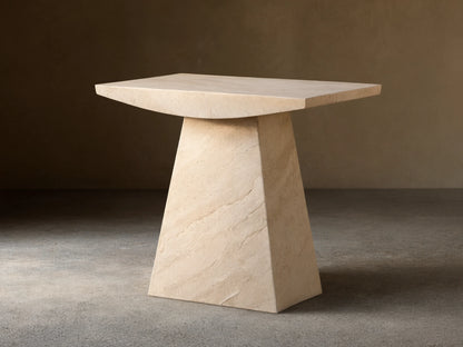 A beige marble side table with a trapezoidal base and a rectangular top, set on a wooden floor against a light wall.