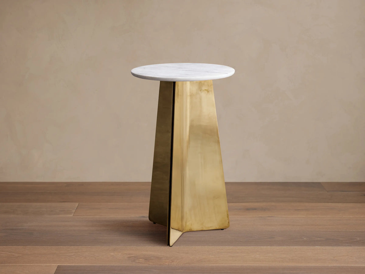 Jorah Martini Table with round white marble top and angular gold metal base, standing on wooden floor against beige wall.
