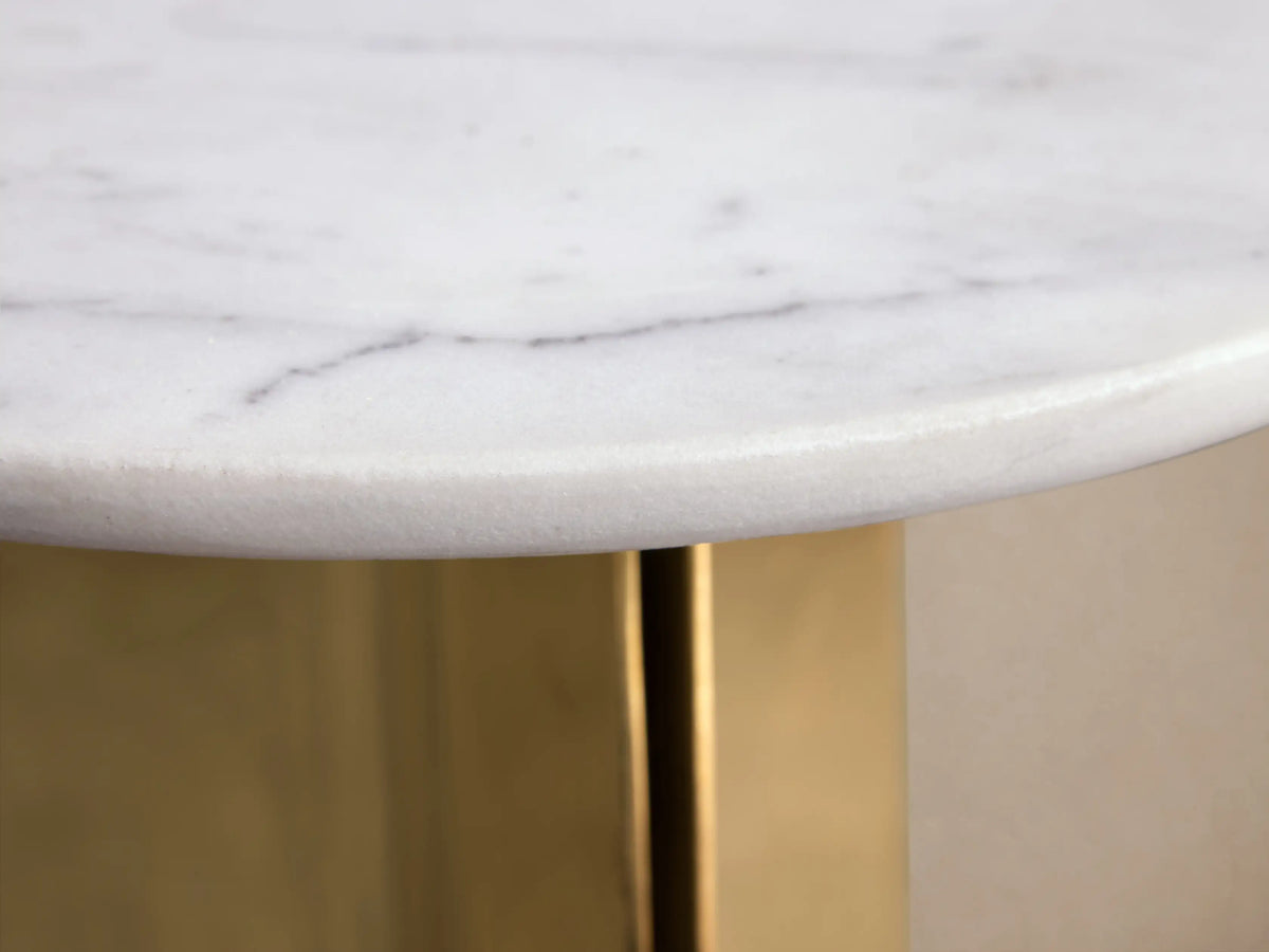 Jorah Martini Table with round white marble top and angular gold metal base, standing on wooden floor against beige wall.