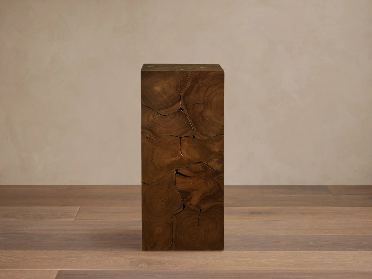 Kalmar Pedestal made of natural teak wood with visible organic shapes, cracks, and patinas. Stands on a wooden floor against a neutral wall.