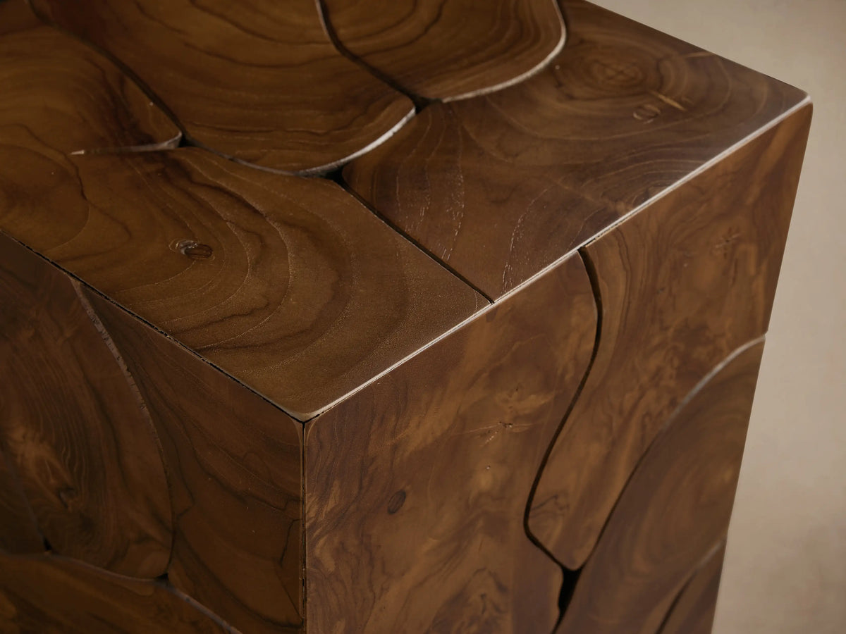 Kalmar Pedestal made of natural teak wood, displaying distinct organic cracks and knots. Set on a light wooden floor against a beige wall.
