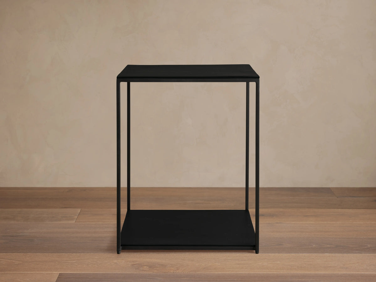 Kavali Square End Table with a black powder-coated finish, solid iron frame, and minimalist design on a wooden floor.