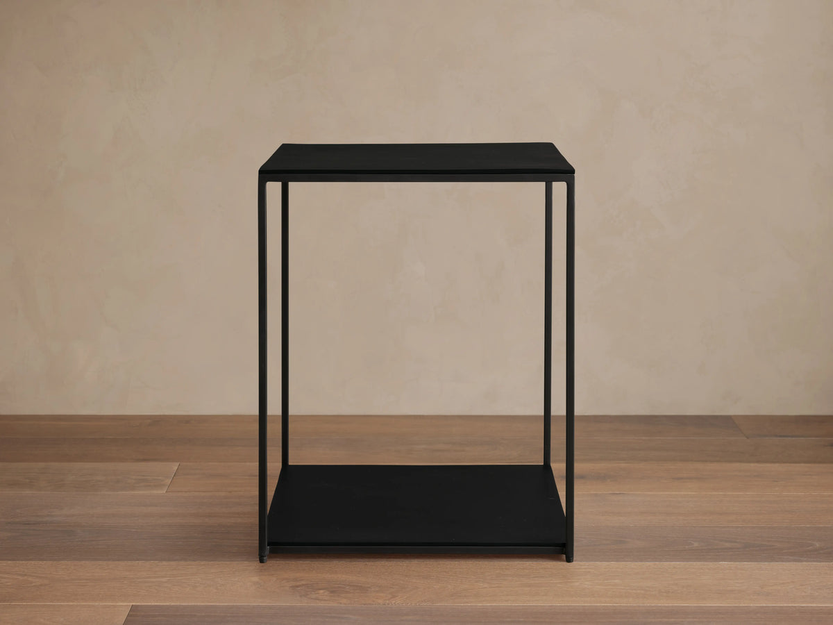 Kavali Square End Table with a black powder-coated finish, solid iron frame, and minimalist design on a wooden floor.