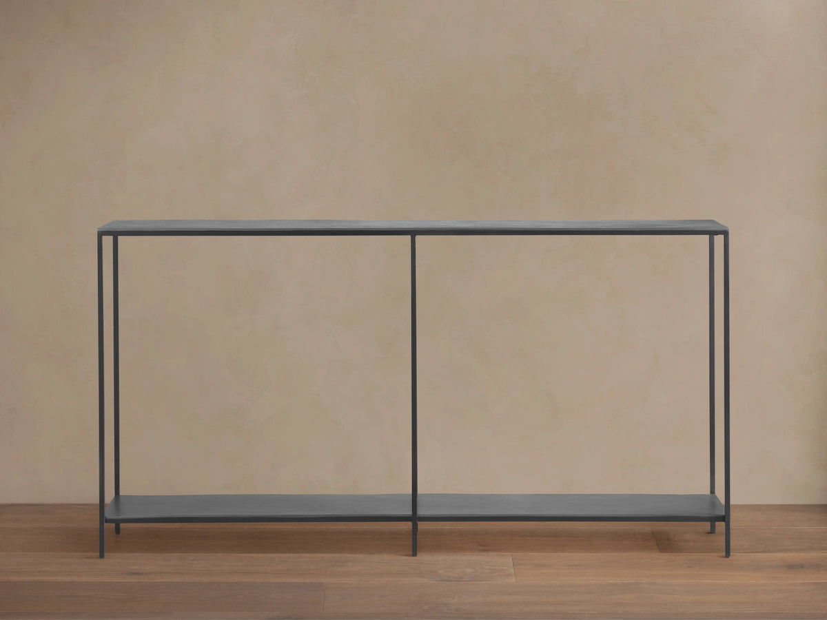 A minimalist Kavali Console Table with a black, powder-coated iron frame, slim design, and two shelves, against a neutral wall.