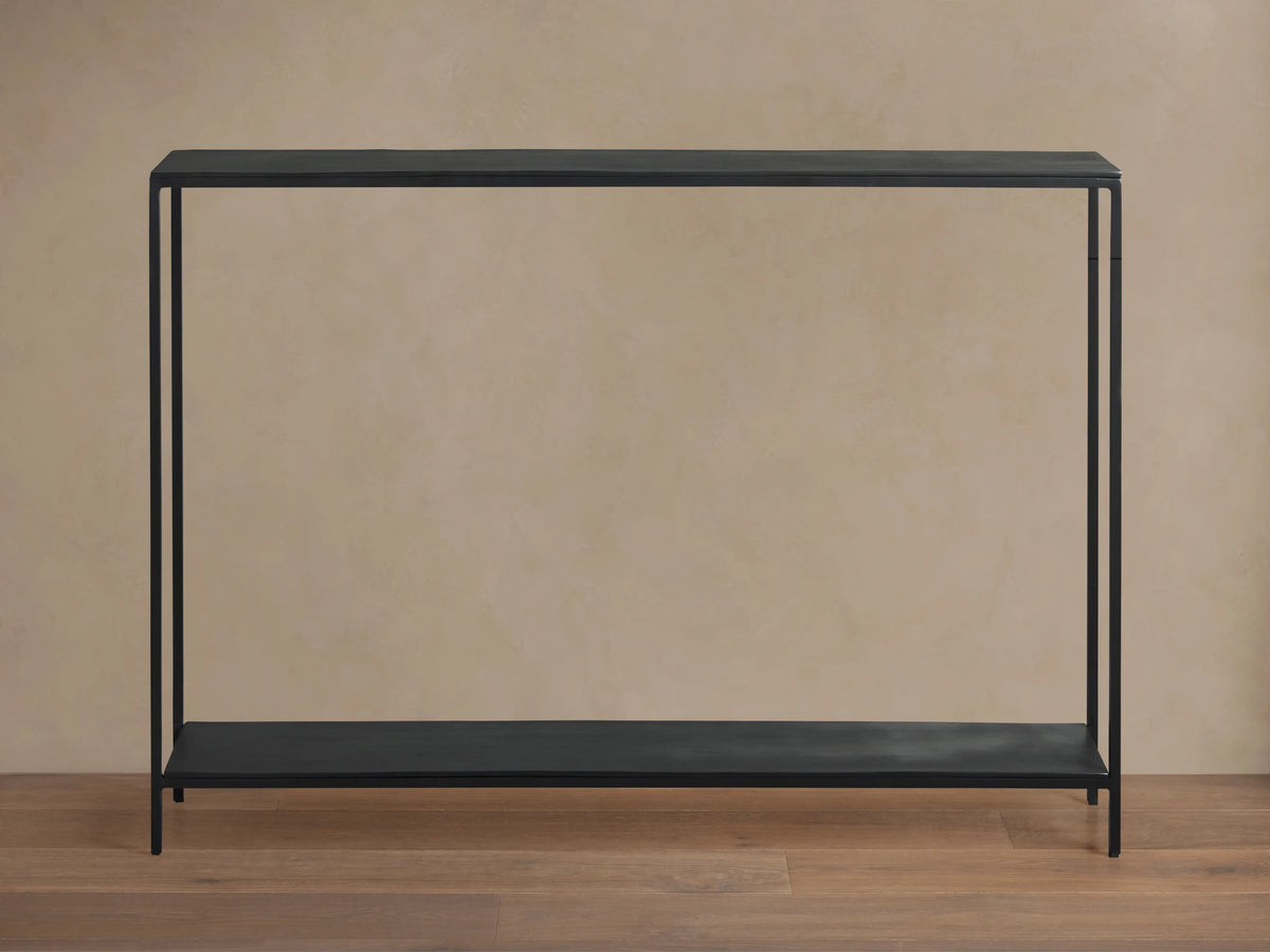 Kavali Slim Console Table, black powder-coated iron frame, rectangular with narrow tabletop and base shelf, on wooden floor.