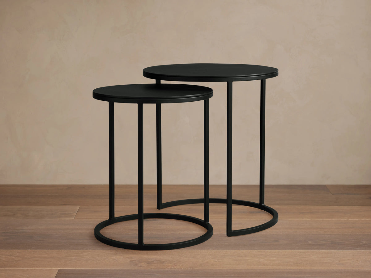 Alt text: Two Kavali Nesting End Tables with round black powder-coated iron frames. The larger table is slightly taller, both set on a wooden floor.