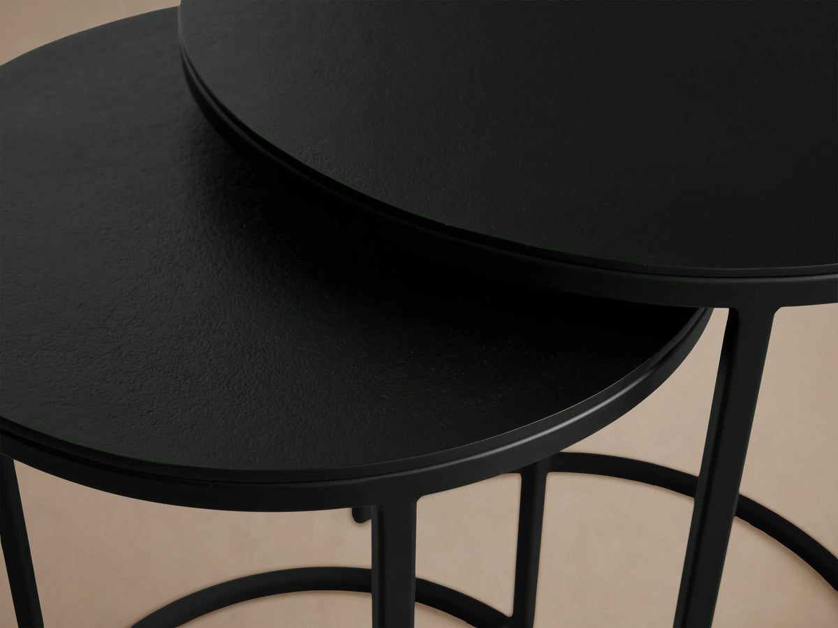 Alt text: Two Kavali Nesting End Tables with round black powder-coated iron frames. The larger table is slightly taller, both set on a wooden floor.