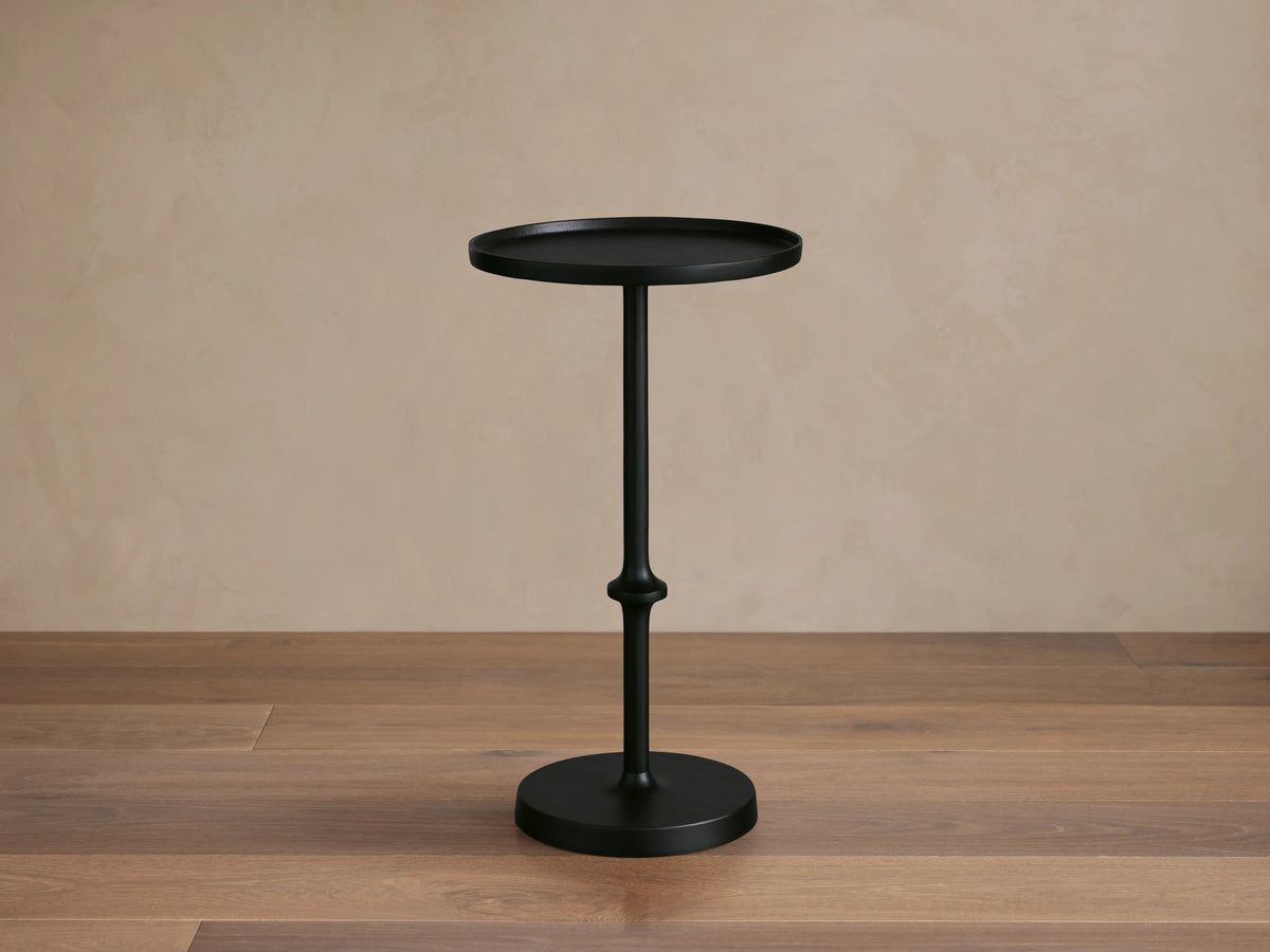 Kenneth Martini Table with a sleek black finish, features a round top and ornate central stem on a wooden floor, set against a beige wall.