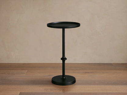 Kenneth Martini Table with a sleek black finish, features a round top and ornate central stem on a wooden floor, set against a beige wall.