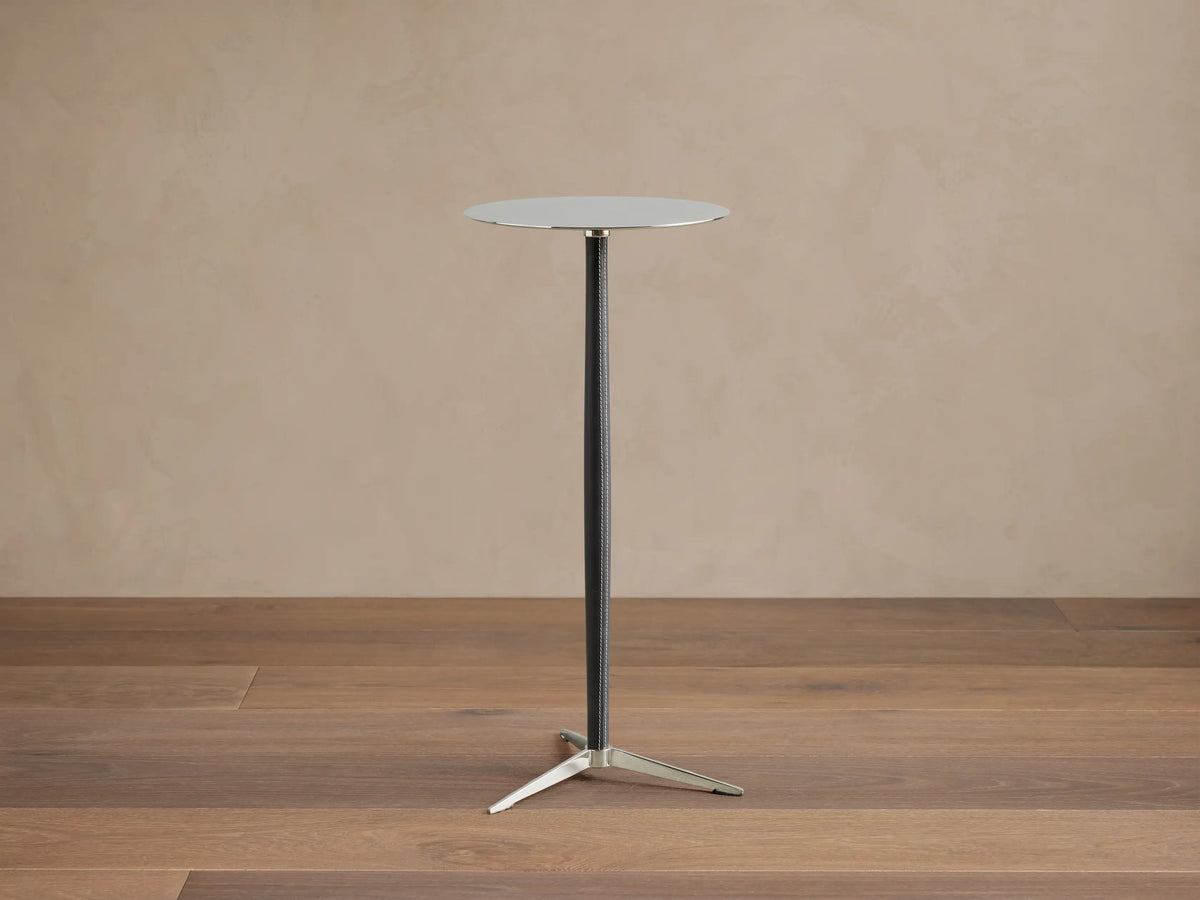 A sleek Lancia Martini Table with a round glass top, slim black leather-wrapped stem, and three-legged stainless steel base.