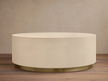 Malone Round Plinth Coffee Table: Cream, round, textured plinth-style coffee table with a golden brass base, on wood floor.