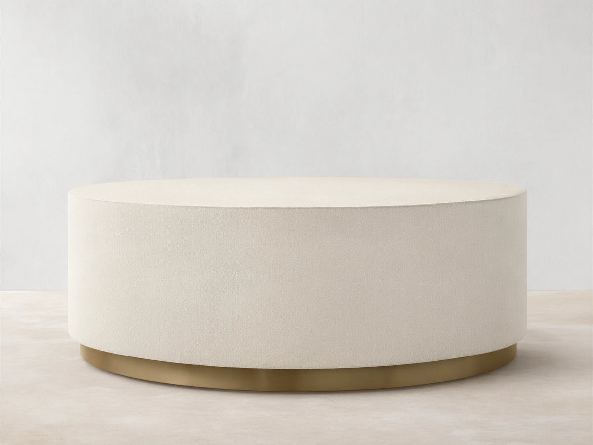 Malone Round Plinth Coffee Table: Cream, round, textured plinth-style coffee table with a golden brass base, on wood floor.