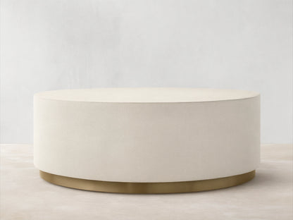 Malone Round Plinth Coffee Table: Cream, round, textured plinth-style coffee table with a golden brass base, on wood floor.