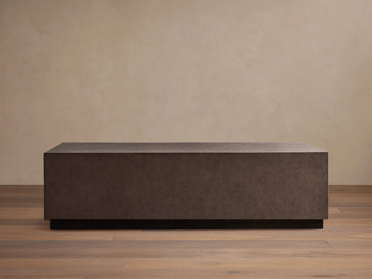 Dark brown rectangular Malone Plinth Coffee Table with textured reconstituted leather finish, on a low dark base, against a beige wall.