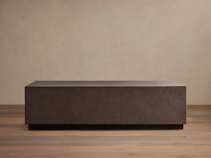 Dark brown rectangular Malone Plinth Coffee Table with textured reconstituted leather finish, on a low dark base, against a beige wall.
