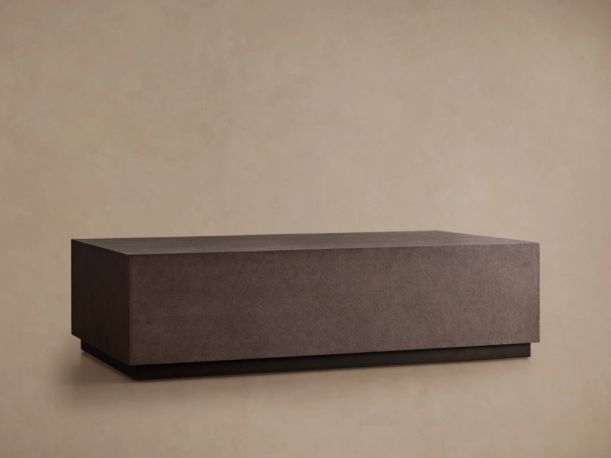 Dark brown rectangular Malone Plinth Coffee Table with textured reconstituted leather finish, on a low dark base, against a beige wall.