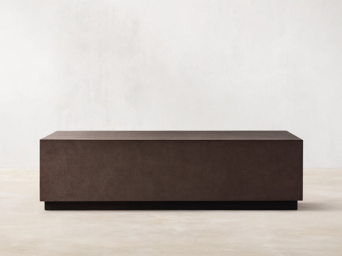 Dark brown rectangular Malone Plinth Coffee Table with textured reconstituted leather finish, on a low dark base, against a beige wall.