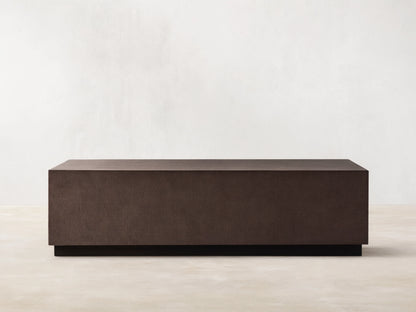 Dark brown rectangular Malone Plinth Coffee Table with textured reconstituted leather finish, on a low dark base, against a beige wall.