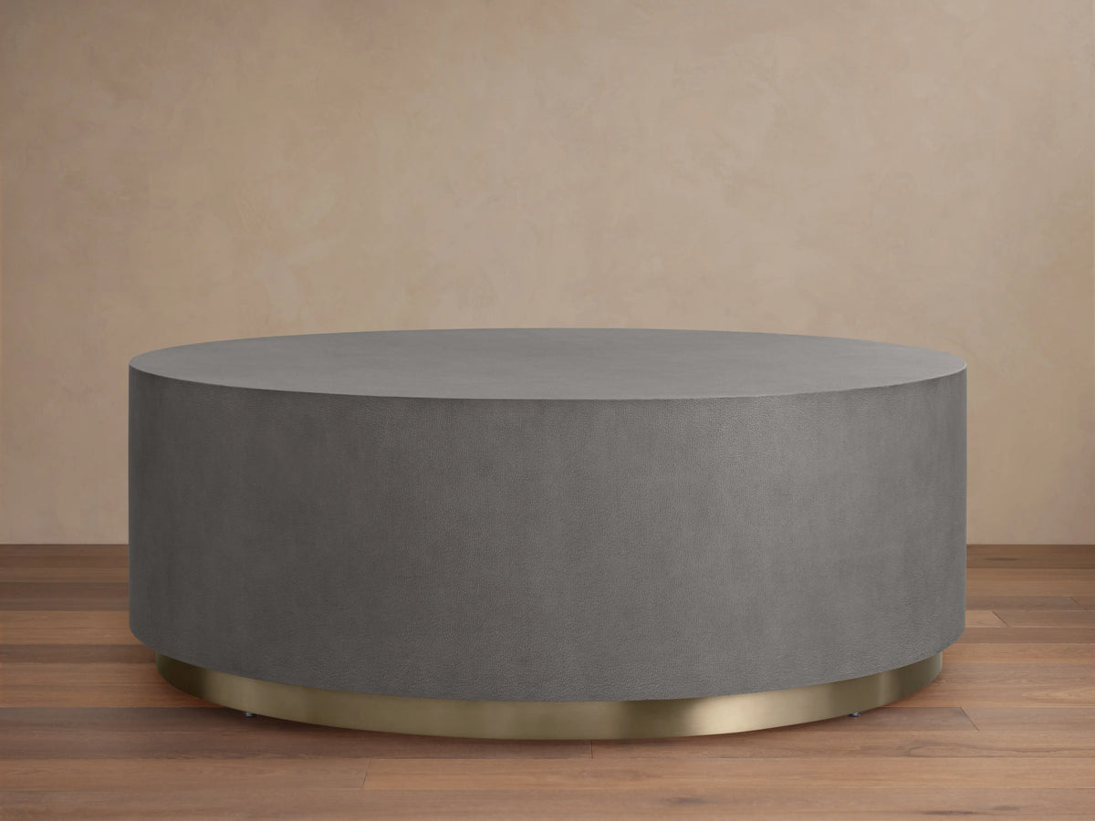Round plinth coffee table with shagreen-patterned grey top, brass-finished steel base. Placed on wooden floor, beige background.