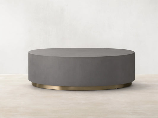 Round plinth coffee table with shagreen-patterned grey top, brass-finished steel base. Placed on wooden floor, beige background.
