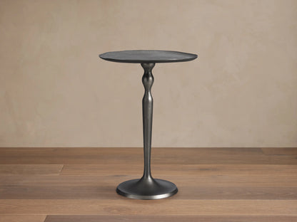 Dark metallic Manhattan Martini Table with textured round top, sculpted stem, and smooth base, on stone floor against beige wall.