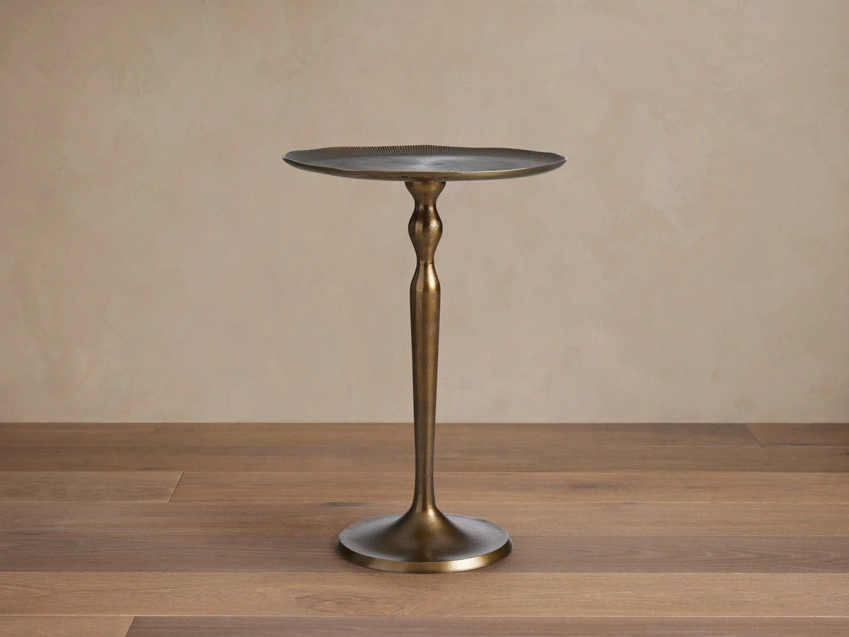 Manhattan Martini Table, dark antique brass finish with a sculpted pedestal base and round textured top, on a tiled floor.