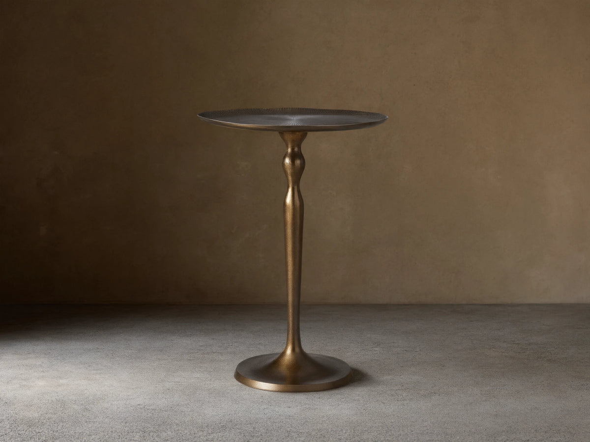 Manhattan Martini Table, dark antique brass finish with a sculpted pedestal base and round textured top, on a tiled floor.