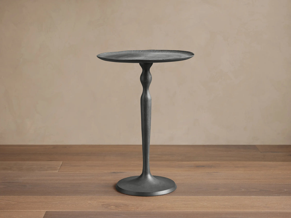 Manhattan Martini Table, dark gray cast aluminum, with round textured top & smooth shapely base, on stone floor against beige wall.