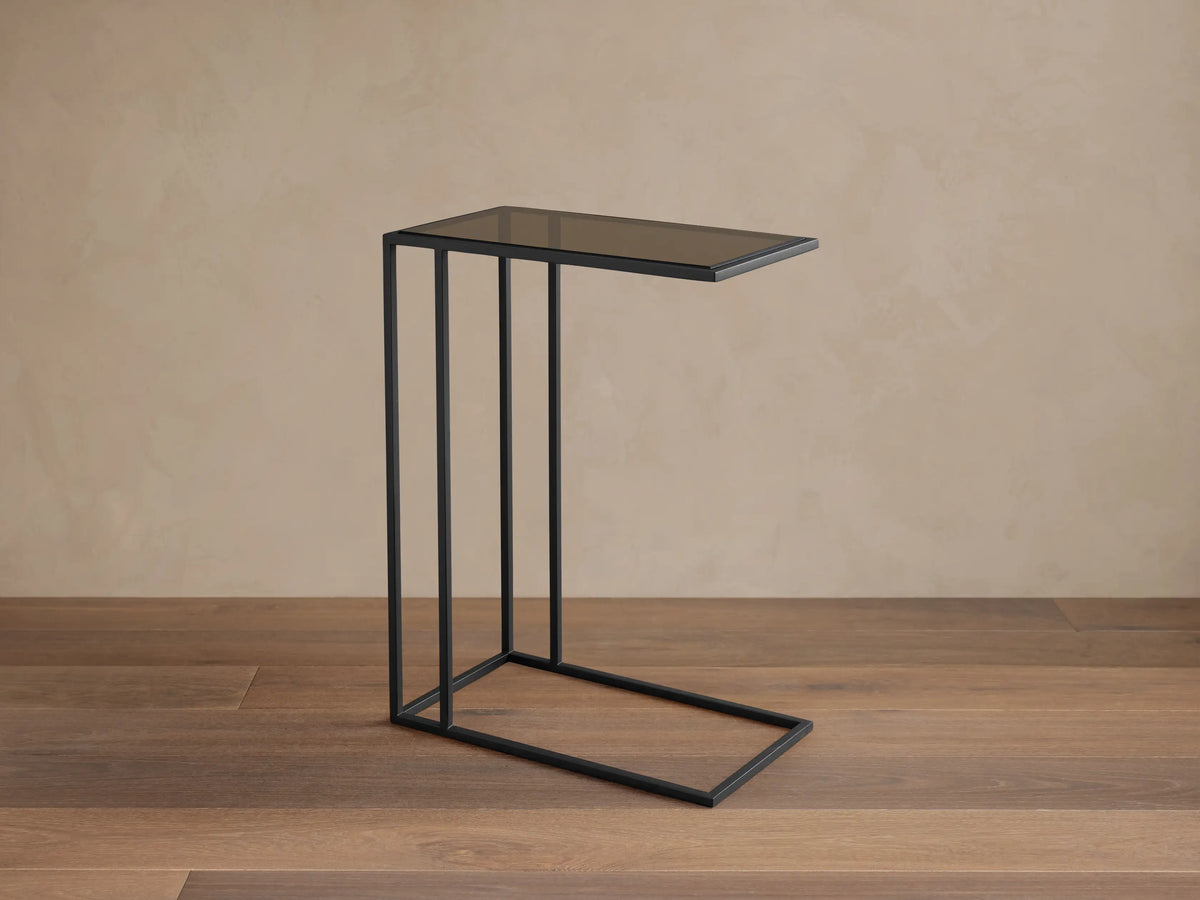 Mills Glass C Table with a matte black steel C-frame and dark glass top, standing on a wood floor against a light wall.