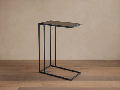 Mills Glass C Table with a matte black steel C-frame and dark glass top, standing on a wood floor against a light wall.