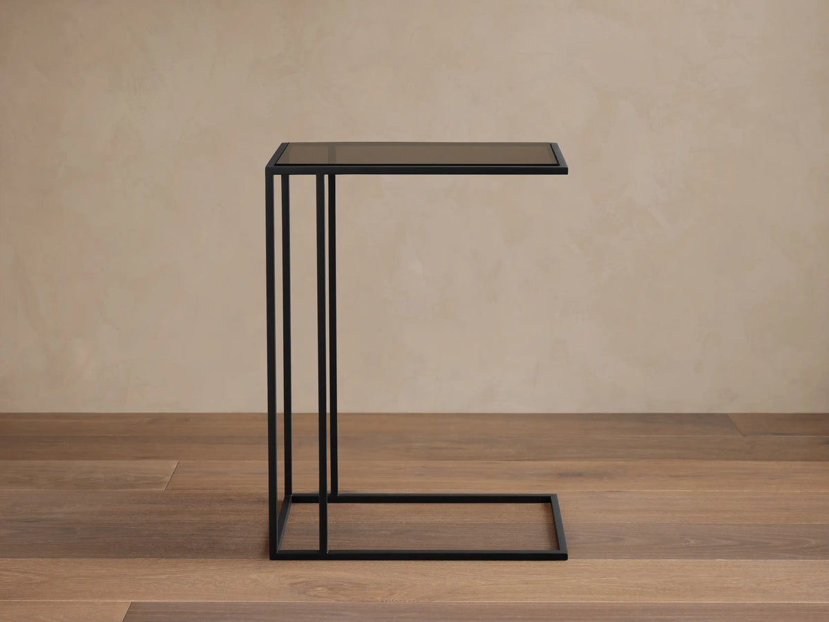 Mills Glass C Table with a matte black steel C-frame and dark glass top, standing on a wood floor against a light wall.