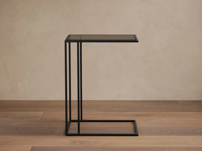 Mills Glass C Table with a matte black steel C-frame and dark glass top, standing on a wood floor against a light wall.